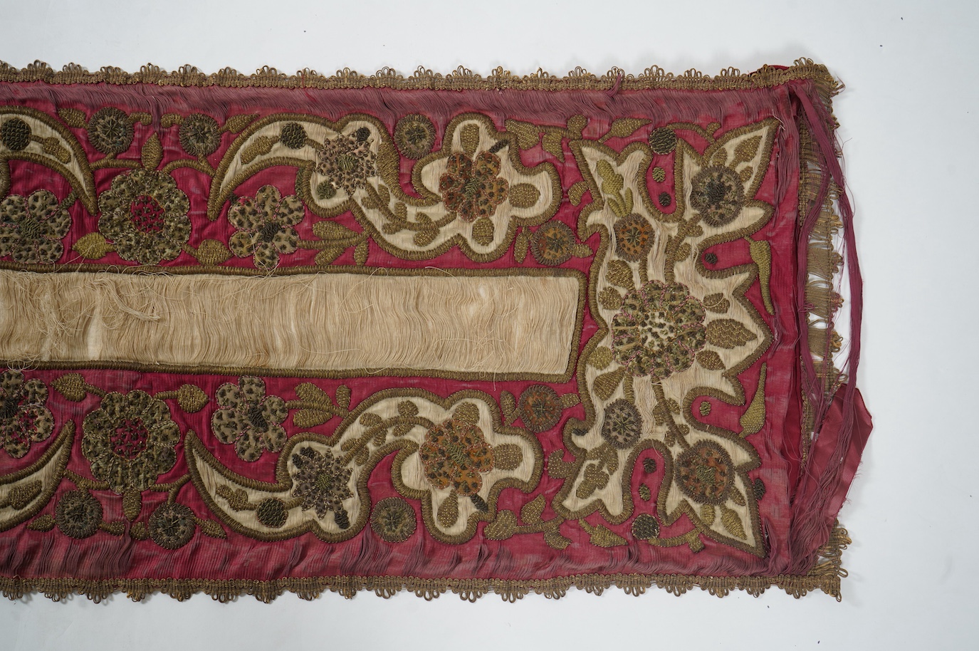 A 19th century Indian red and cream silk runner, heavily embroidered with scrolling motifs, using a variety of stump work and gold wire work stitches, with earlier Mughal influences, 118cm x 46.5cm (not including braid),